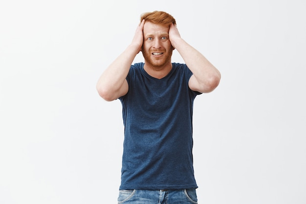 Portrait of man in despair feeling regret, standing anxious over gray wall, clenching teeth, holding hands on head and frowning