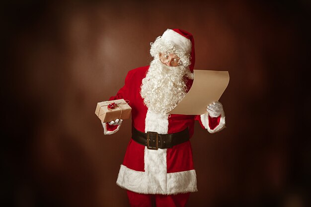 Portrait of Man in Santa Claus Costume - with a Luxurious White Beard, Santa's Hat and a Red Costume reading letter at red studio background with gifts