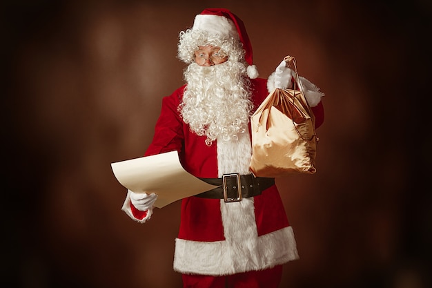 Free photo portrait of man in santa claus costume - with a luxurious white beard, santa's hat and a red costume reading letter at red studio background with gifts