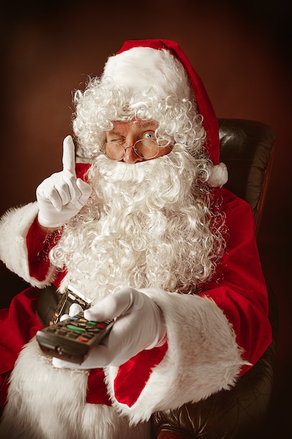 Free photo portrait of man in santa claus costume with a luxurious white beard, santa's hat and a red costume at red sitting in a chair with tv remote control