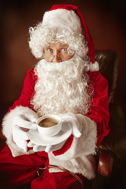 Portrait of Man in Santa Claus Costume - with a Luxurious White Beard, Santa's Hat and a Red Costume at red studio background sitting in a chair with cup of coffee