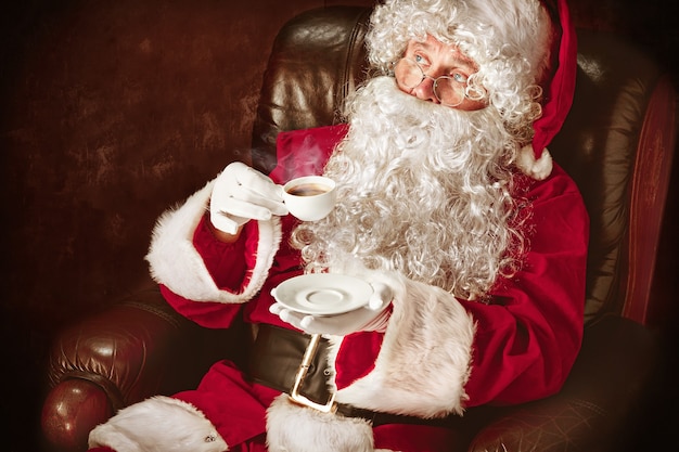 Free photo portrait of man in santa claus costume - with a luxurious white beard, santa's hat and a red costume at red studio background sitting in a chair with cup of coffee