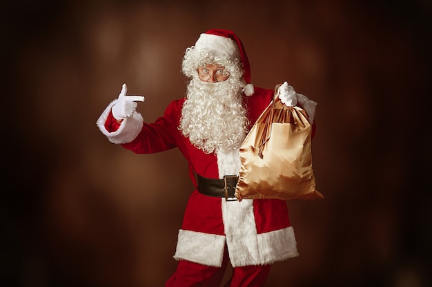 Free Photo portrait of man in santa claus costume - with a luxurious white beard, santa's hat and a red costume at red studio background with gifts