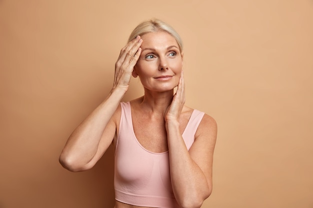 Free photo portrait of mature elderly european woman touches face gently has perfect skin and looks thoughtfully away enjoys her soft complexion cares about appearance satisfied after anti aging procedure