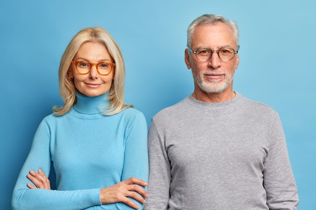 Portrait of mature woman and man stand next to each other in casual clothes against blue wall look directly at front with calm expressions