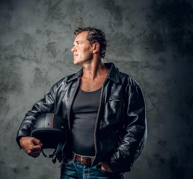 Free Photo portrait of a middle age male dressed in a leather jacket holds motorcycle helmet over grey background.