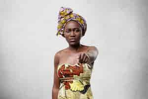 Free photo portrait of middle-aged black woman with slender figure wearing scarf on head and beautiful dress standing against white concrete wall pointing at you with forefinger having serious expression