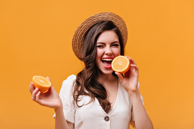 Portrait of mischievous girl with wavy hair biting orange. Lady in straw hat posing on orange background.