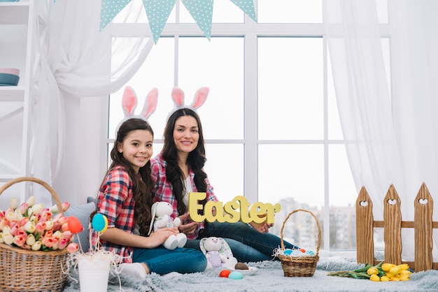 Free Photo portrait of mother and daughter sitting near the window holding easter word and bunny in hand