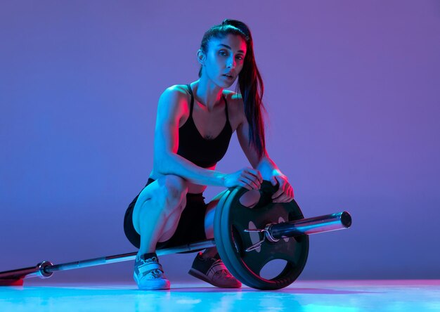 Portrait of muscled woman training with a barbell isolated on purple background in neon light