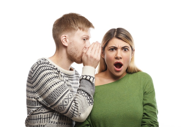 Free photo portrait of mysterious young fair-haired male with stubble covering ear of his shocked girlfriend who is listening to gossips, rumours, confidential information and secrets with eyes full of disbelief