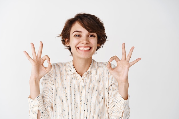 Free Photo portrait of natural happy woman with short hairstyle, showing okay gestures and smiling, approve and like something, show positive feedback, white wall