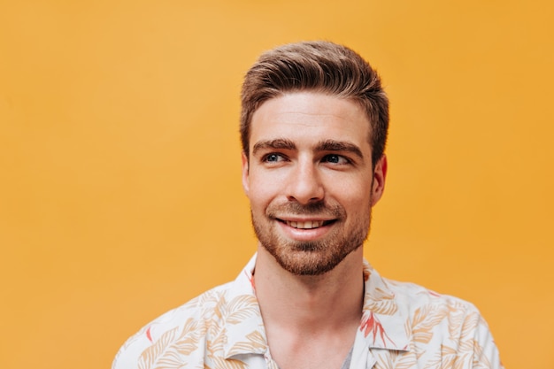 Portrait of nice young man with stylish hairstyle, blue eyes and beard in light cool shirt looking away and smiling on orange wall