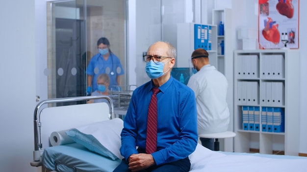 Free photo portrait of old man wearing mask at doctor appointment, sitting on the hospital bed waiting for covid-19 results. healthcare mecical medicine system during global pandemic, handheld slow motion shot