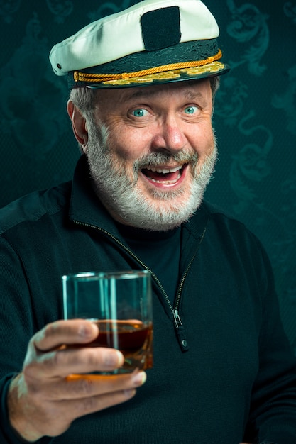 Free photo portrait of old sailor man as captain in black sweater and hat drinking cognac on black studio