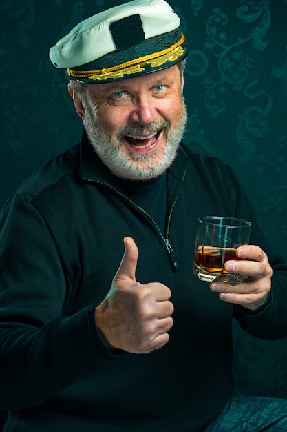 Free Photo portrait of old sailor man as captain in black sweater and hat drinking cognac on black studio