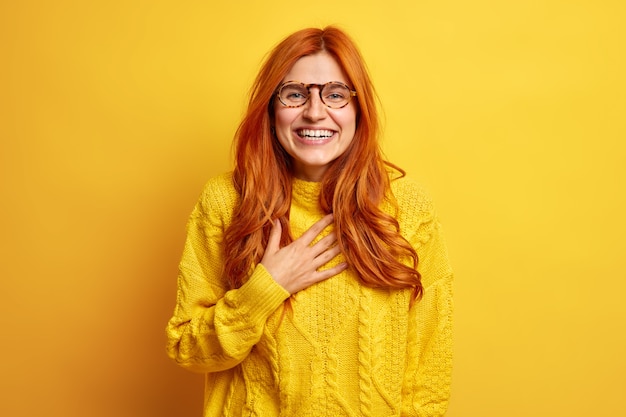 Portrait of overjoyed redhead woman smiles broadly keeps hand on chest smiles broadly expresses good emotions feels pleased wears transparent glasses casual sweater.