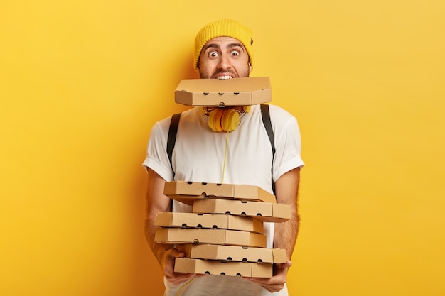 Portrait of overloaded pizza man holds many cardboard packages, one in mouth, wears casual white t shirt.