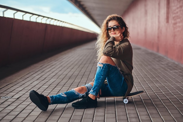 Free photo portrait of a pensive girl in sunglasses dressed in a hoodie and ripped jeans sitting on a skateboard at a bridge footway.