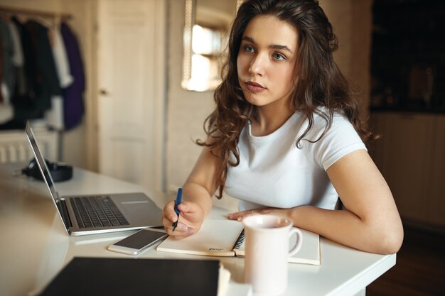 Portrait of pensive thoughtful student girl studying from home, sitting at her workplace in front of open laptop