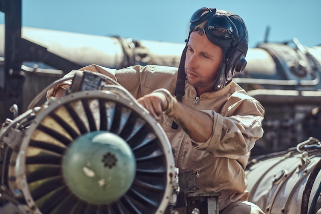 Free Photo portrait of a pilot-mechanic in uniform and flying helmet, repairing the dismantled airplane turbine in an open-air museum.