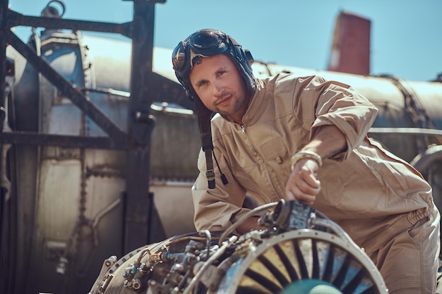 Free photo portrait of a pilot-mechanic in uniform and flying helmet, repairing the dismantled airplane turbine in an open-air museum.