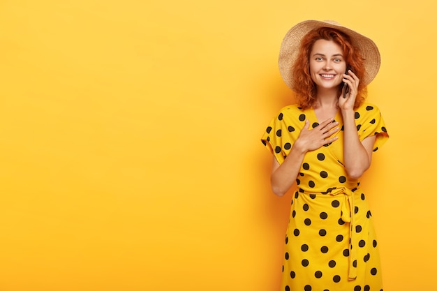 Free photo portrait of pleasant looking satisfied young redhead woman keeps palms on chest, feels impressed to hear heart piercing story over smartphone, wears stylish bright yellow summer outfit