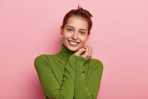 Free Photo portrait of pleasant looking young woman with toothy smile, keeps hands near cheek, dressed in casual poloneck, gazes happily, enjoys good news, poses against pink background.