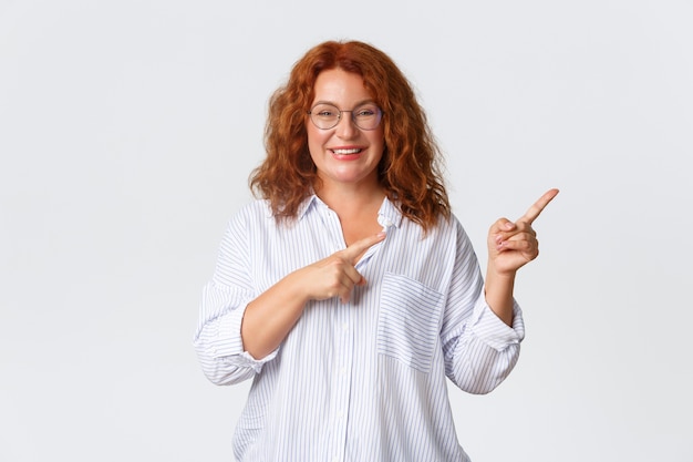 Portrait of pleasant smiling middle-aged woman with red hair, wearing glasses and blouse showing advertisement, client of company recommend product or service, pointing right.