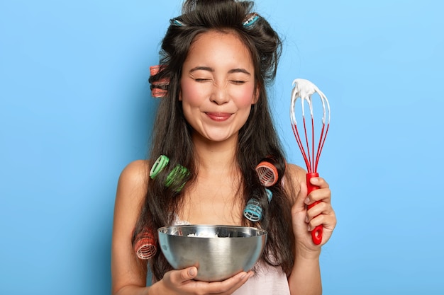 Free photo portrait of pleased beautiful korean lady holds beater and bowl, makes sweet cream for biscuit cake, wears hair curlers