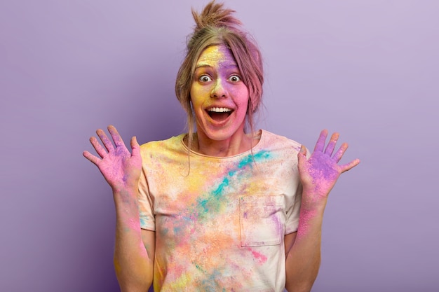 Portrait of pleased female has colored face, shows palms smeared with powder, expresses happiness and surprised reaction, poses against purple wall, celebrates traditional Indian holiday