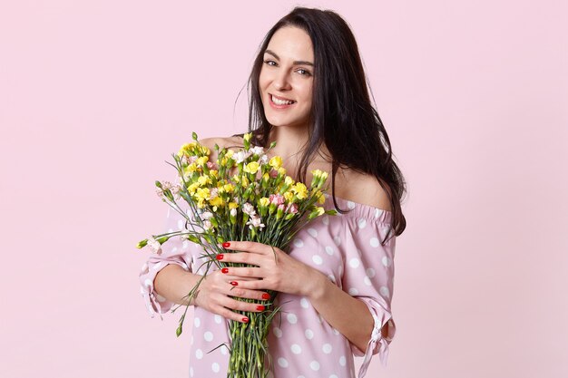 Portrait of pleased female model with dark hair, dressed in polka dot dress, smiles positively