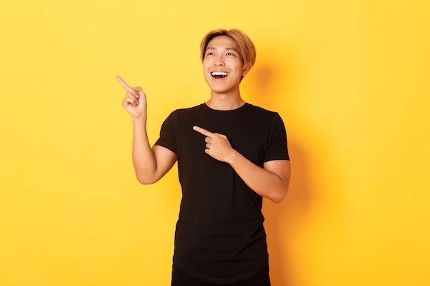 Portrait of pleased handsome asian guy in black t-shirt, pointing fingers and looking upper left corner with satisfied smile, yellow wall