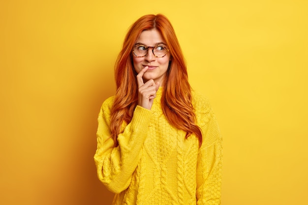 Portrait of pleased nice looking young woman keeps index finger near lips concentrated aside has thoughtful expression has natural red hair wears casual sweater.