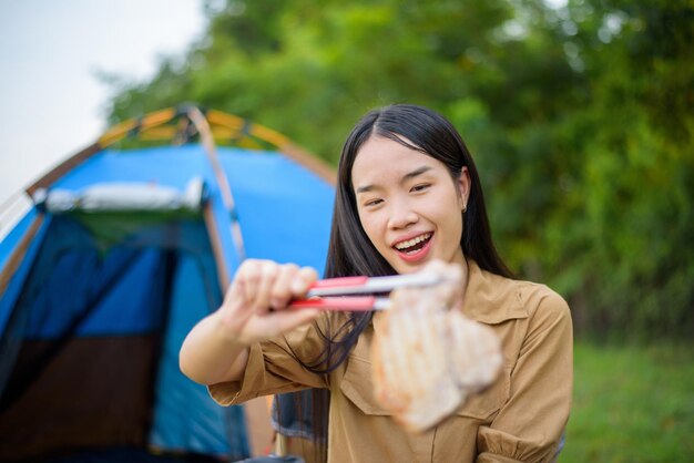 Portrait of Portrait of happy young asian woman camping alone grilled pork barbecue in the picnic pan and cooking food while sitting on chair in the camping site