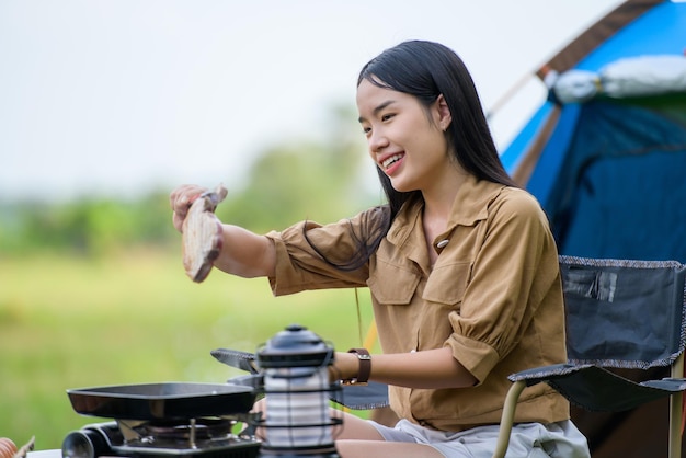 Portrait of Portrait of happy young asian woman camping alone grilled pork barbecue in the picnic pan and cooking food while sitting on chair in the camping site