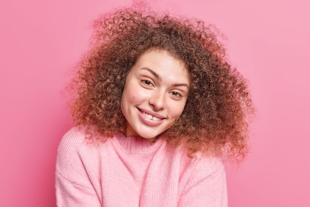 Free photo portrait of positive alluring woman with curly bushy hair smiles gently  shows white teeth has healthy skin wears casual jumper isolated over pink wall. natural beauty concept