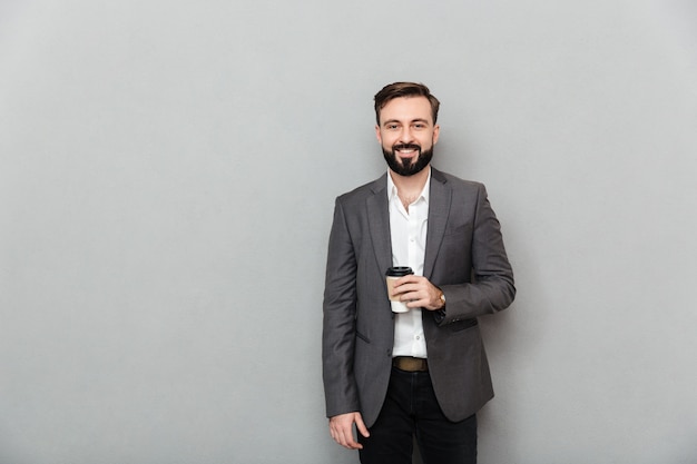 Portrait of positive man in white shirt posing on camera with broad smile, and holding takeaway coffee over gray