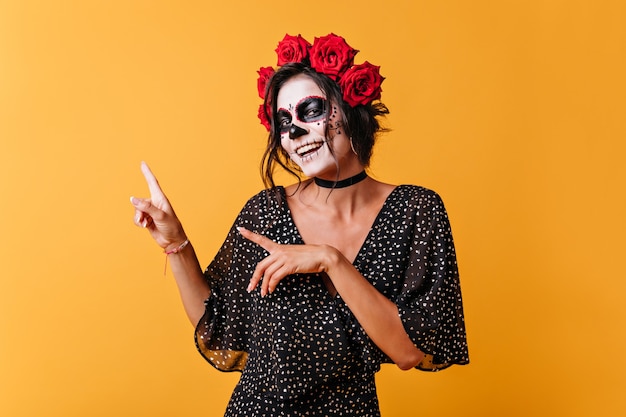 Portrait of positive Mexican girl on orange background with space for text. Woman with skull mask smiles cutely and points fingers up.