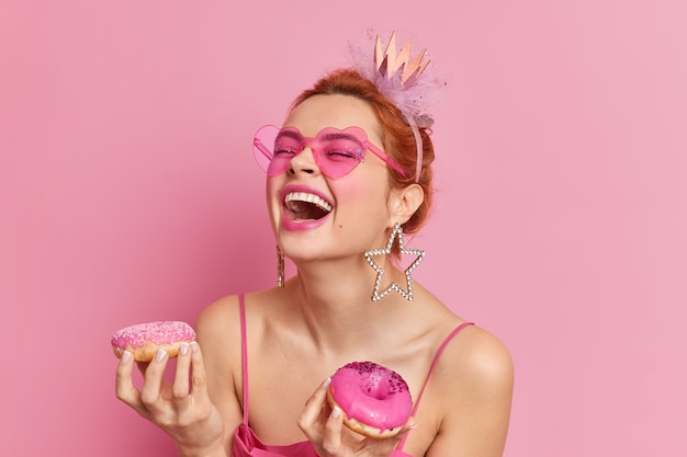 Free photo portrait of positive redhead caucasian woman laughs loudly keeps mouth opened has upbeat mood holds two doughnuts