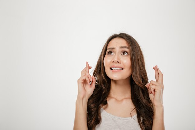 Portrait of praying young woman wearing casual begging god please looking up with keeping fingers crossed, isolated over white