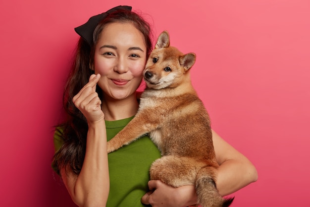 Portrait of pretty Asian woman shows mini heart hand gesture, expresses love, embraces Japanese pedigree hunting dog, enjoys spending free time with favorite pet