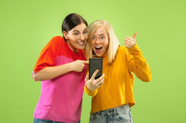 Portrait of pretty charming girls in casual outfits isolated on green studio background. Girlfriends or lesbians making selfie. Concept of LGBT, equality, human emotions, love, relation.