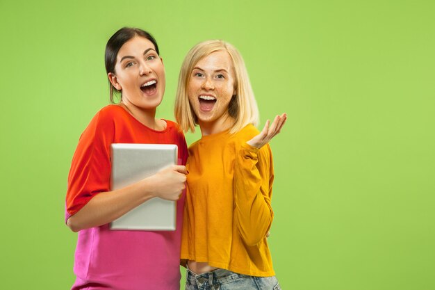 Portrait of pretty charming girls in casual outfits isolated on green studio background. Girlfriends or lesbians using a tablet for fun or payments. Concept of LGBT, human emotions, love, relation.