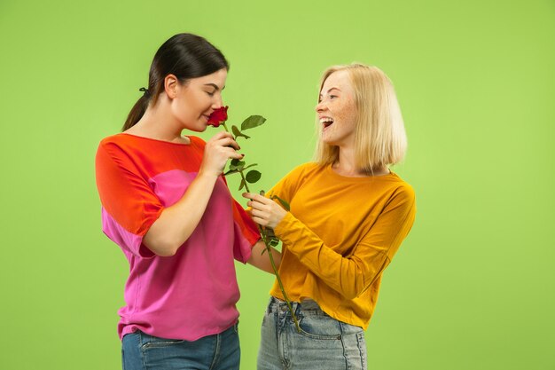 Portrait of pretty charming girls in casual outfits isolated on green studio background. Two female models as a girlfriends or lesbians. Concept of LGBT, equality, human emotions, love, relation.