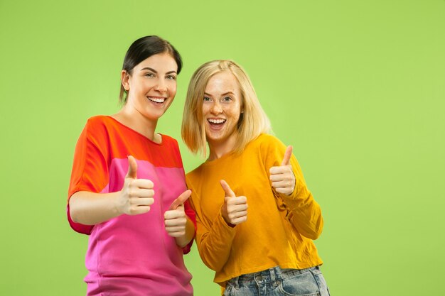 Portrait of pretty charming girls in casual outfits isolated on green studio background. Two female models as a girlfriends or lesbians. Concept of LGBT, equality, human emotions, love, relation.