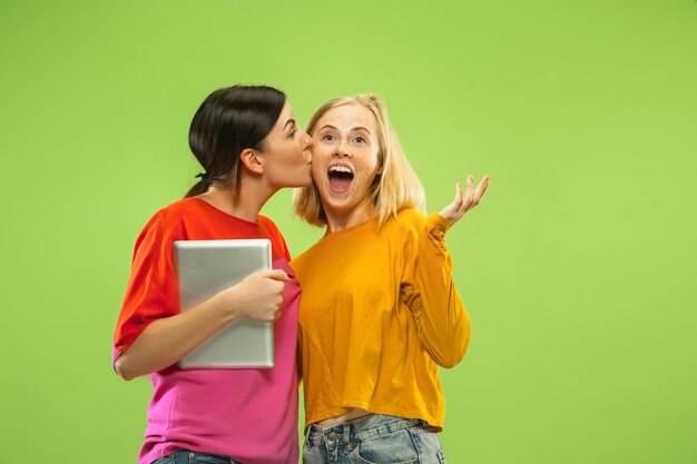 Portrait of pretty charming girls in casual outfits isolated on green wall. Girlfriends or lesbians using a tablet for fun or payments. Concept of LGBT, human emotions, love, relation.