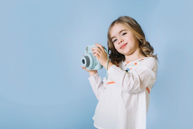 Portrait of a pretty girl holding retro instant camera in hands against blue background