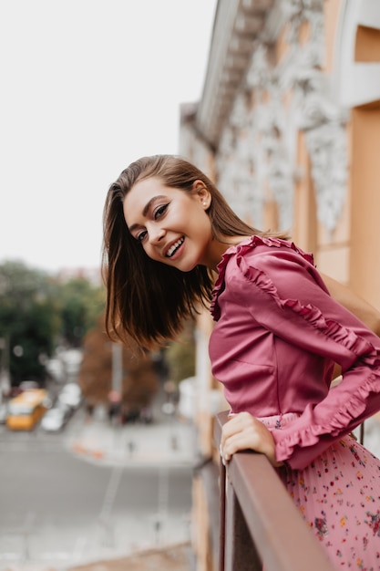 Portrait of pretty girl peeking out from behind balcony railing. Young traveler with short hair is happy that she sees new old city