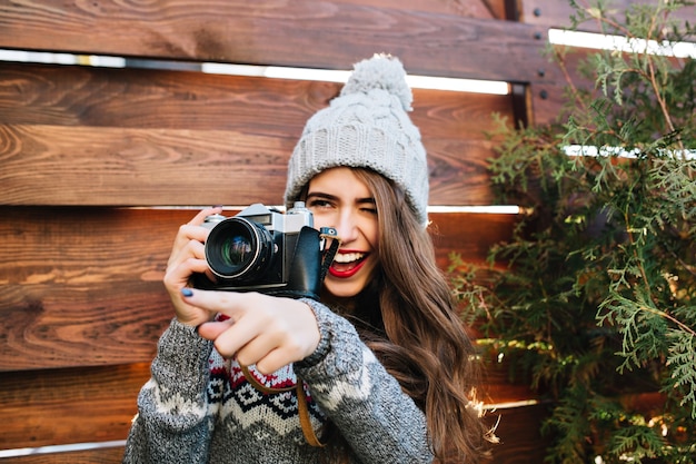 Free photo portrait pretty girl  with long hair in knitted hat having fun at making a photo on camera on wooden .